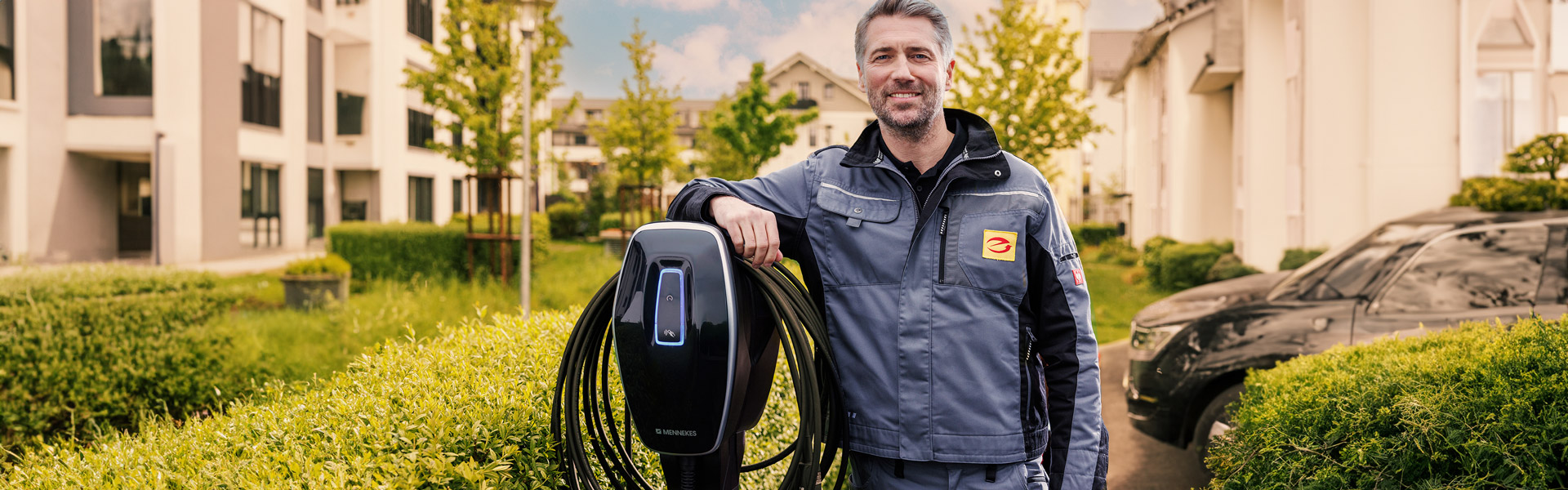 Installateur stützt sich auf Wallbox ab, die auf einer Stehle auf einem Parkplatz steht