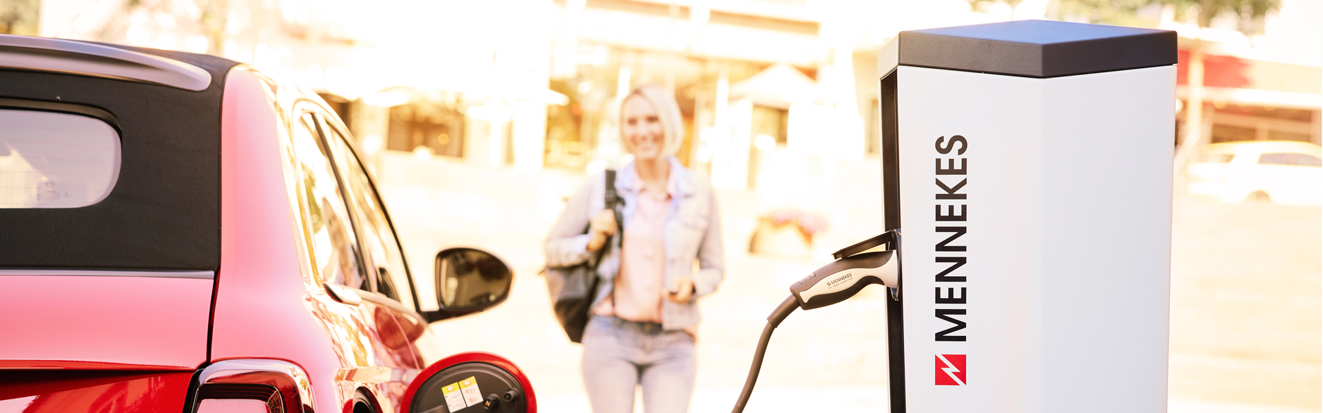 E-Auto lädt an einer öffentlichen Ladesäule, Frau kommt auf das Auto zu