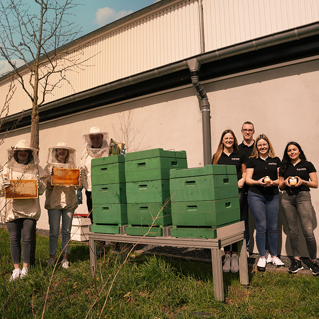 Gruppe von Menschen, die neben mehreren grünen Bienenstöcken stehen; auf der linken Seite tragen einige Personen Imkeranzüge und halten Wabenrahmen