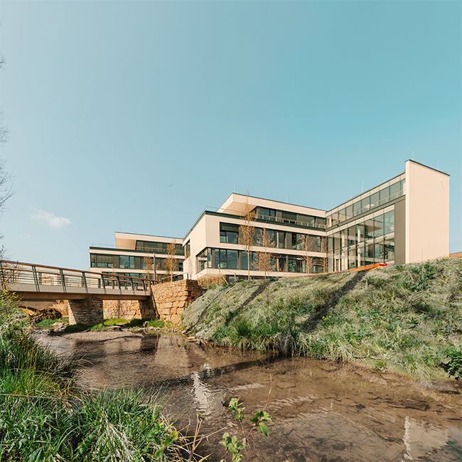 Modernes MENNEKES-Gebäude mit großen Fenstern und einer Brücke im Vordergrund, über einen kleinen Bach führend.