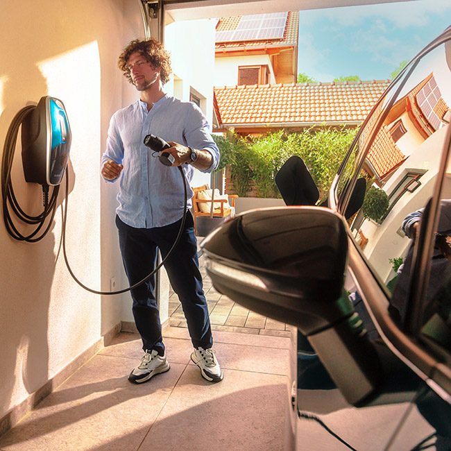 Mann, der ein Elektroauto in einer Garage auflädt; im Hintergrund ist ein modernes Haus mit Solarpanelen auf dem Dach zu sehen