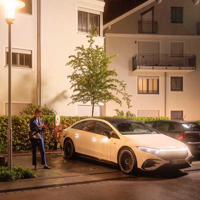 Eine Frau lädt nachts ihren Dienstwagen an einer Wallbox vor einem modernen Wohnhaus auf.