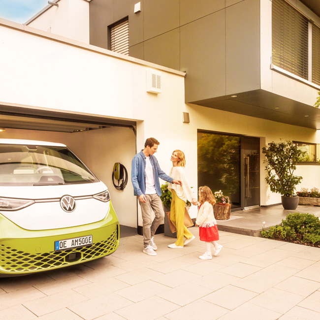 Eltern mit Tochter vor dem Haus mit einer Garage, in welcher ein E-Auto und eine Wallbox hängt