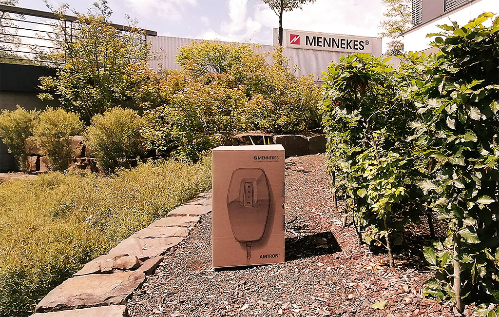 Ein MENNEKES-Karton steht auf einem gepflasterten Weg in einem Gartenbereich, im Hintergrund ist ein MENNEKES-Gebäude zu sehen