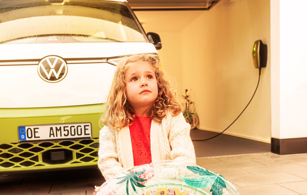 Ein kleines Mädchen sitzt vor einem in der Garage geparkten Volkswagen-Elektroauto, das an eine Ladestation angeschlossen ist