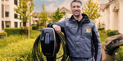 Installateur stützt sich auf Wallbox ab, die auf einer Stehle auf einem Parkplatz steht